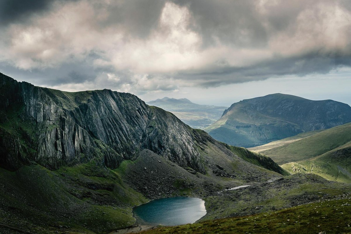 A broad mountain valley with a small lake below dramatic ridges and clouds.